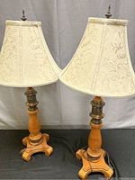 Photo of both lamps side by side showing full size, cream linen shades with embossed floral patterns, turned wooden bases and brass accents, sitting on black surface.