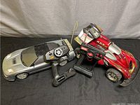 Two remote control cars, one silver sports car and one red off-road car with lightning bolt decals and number 22, each with their respective remote controllers, laid on a dark surface.