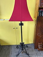 Full view of floor lamp showing bent iron base with curled decorative details and red fabric shade against yellow background.