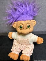 Front view of purple haired troll doll wearing striped overalls and white shirt, green eyes, orange hands and feet.