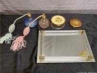 Two vintage perfume spritzers, round powder container, compact, and rectangular mirror tray on a black cloth background.