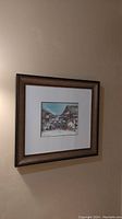 Framed watercolour painting hanging on a beige wall, showing street scene with buildings and clear sky.