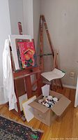 Corner view showing two larger wooden easels, one small tripod easel, metal stool, cardboard box of paints and a painting pad on the floor
