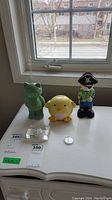 View of three coin banks (green frog, yellow chick, pirate) along with two coins placed on white furniture near window.