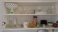 Open white cabinet showing a variety of glassware items including milk glass compote, teacups, and other glasses arranged on shelves.