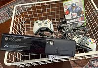 Xbox 360 E console, two controllers, cables, and Madden NFL 07 game all placed inside a white wire basket on wooden floor.