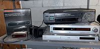Shelf with 4 electronic devices: Sony VCR on top, Sony DVD changer, Malata and Sylvania DVD players on lower shelf. Box for Sylvania DVD player on left.