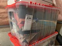Clear plastic storage bin filled with assorted fabrics, red snap-on lid visible
