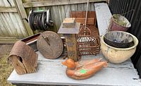 Overview of garden decor items placed on outdoor table including wooden birdhouse, metal sundial, wood duck sculpture, metal cage, and several plant pots.