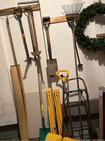 Photo showing various garden tools hanging on a wall including shovels, rakes, snow shovel, hand truck, and a rolled-up mat.
