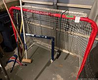 Wide view of large collapsible Rawlings hockey net with red metal frame and white netting with smaller blue plastic framed net in front. Several hockey sticks and worn curling broom leaning against nets.