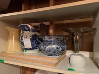 Shelf with two ceramic pitchers, one blue and white and one off-white, a blue and white ceramic bowl, a silver plate vase, and a small white ceramic creamer.