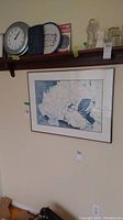 Photo of framed watercolour print by V May and items on wooden wall shelf above, includes clock, plate, plaque, glass figurine, glasses, and bottle.