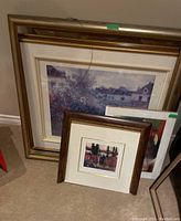 Four framed prints leaning against wall. One large floral print in wood frame, smaller prints in front including Tom Thomson reproduction in broken frame.