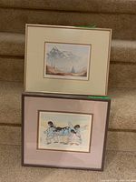Two metal framed art prints stacked on carpeted stairs. Top print features mountain landscape. Bottom print features Inuit dancers in traditional winter clothing.