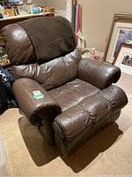 Brown faux leather recliner armchair viewed at a slight angle showing padded rolled arms, seat, and backrest with fabric cover on headrest.