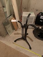 Full view of black cast iron table base standing on basement floor near a mirror and dehumidifier, showing overall shape and height with measuring tape extended horizontally at base.