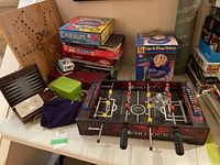 Photo showing Pro Kick foosball game with multiple games stacked behind including Cranium and bingo set, plus various smaller game accessories on table.