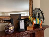 Photo featuring ceramic jar, black metal cow bell, colorful wooden horse figurine, brass vase and framed photo in background on shelf