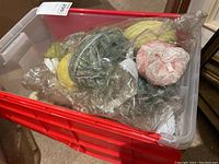 Top view of plastic bin containing various balls of yarn, some wrapped in plastic layers, with visible colors including yellow, green, pink, and beige.