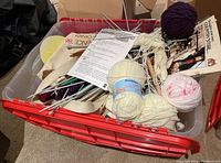 Overall view of the plastic bin with yarn skeins, knitting needles, and booklets inside.
