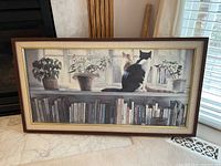 Full view of the framed art print depicting two cats on a windowsill with surrounding plants and books below.
