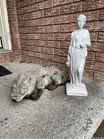 Front view of a weathered gray cement dragon statue beside a white hollow stone-like classical lady statue on a porch step