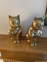 Front view of two brass cat figurines placed side by side on a wooden surface showing detailed faces and polished brass finish.