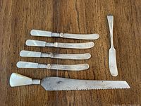 Full view of six silver-plated butter knives and a bread knife with mother-of-pearl handles, laid out on a wooden surface.