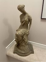 Front view of Aphrodite plaster statue seated with a draped garment, placed against wall corner on a carpeted floor.