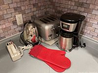 Shows the full set of kitchen appliances on a countertop: kettle, toaster, coffee maker, hand mixer and oven mitts.