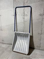 Full view of large snow shovel leaning against a concrete wall showing white plastic scoop and blue metal handle
