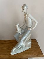 Front and side view of porcelain figurine depicting a young man sitting on a rock with a dog, showing delicate features and smooth glossy finish.