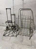 Photo of a metal wire basket with handle and wheels next to a foldable metal trolley with telescoping handle, both on a concrete floor against a concrete wall.