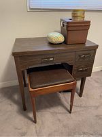 Sewing table with stool, showing closed table top and items on top: pincushion and brown storage box.