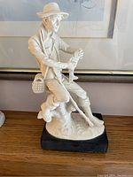 Full view of the white ceramic figurine of a seated fisherman on a black base, holding fish in hands, wearing a hat and fishing basket at side.