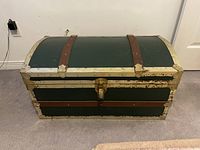 Closed view of trunk showing dark green paint, off-white edges, brown leather straps, and brass latch.