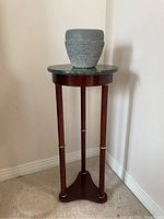 Wooden planter stand with green stone top and gray ceramic planter on top, standing on carpeted floor near wall