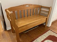 Overall view of wooden storage bench showing slatted backrest, armrests, and storage compartment under hinged seat. Bench placed against a white wall on wood flooring next to a patterned area rug.