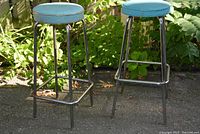 Two vintage 1960s metal-legged bar stools with blue round cushioned seats, showing used condition and outdoor setting.