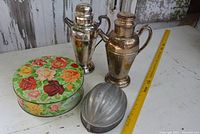 Photo showing all items together: 2 silver-tone cocktail shakers, floral Peak Frean cookie tin, and 2 metal food molds on a white surface with yellow measuring stick.