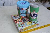 Three puzzles displayed: blue tin labeled 'Books', green tin labeled 'Beer Lovers', and flat puzzle box with New York City life scene showing people, bicycle, yellow taxi, dog, cafe, and street elements.