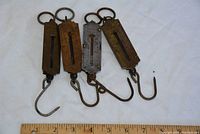 Four vintage fish weight scales with metal hooks and rings laid out horizontally, next to a ruler for size reference.