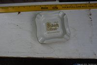 Top view of the square white ceramic ashtray with gold lettering "Asbach Uralt" and a gold trim around the edges, on a white surface with a measuring tape indicating size.