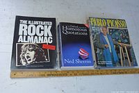 Three books lined up on a white surface with a ruler for scale: The Illustrated Rock Almanac, The Oxford Book of Humorous Quotations, Pablo Picasso.