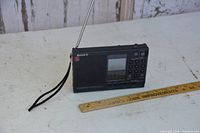 Full front view of Sony ICF-SW7600 radio with antenna extended and wrist strap visible, ruler showing approximate size.