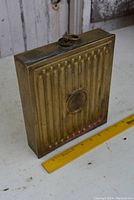 Side view of rectangular brass canteen with vertical ridges and brass cap, placed next to a yellow ruler showing approx. 8.5 inches in height.