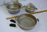 Three vintage aluminum cooking pieces including two pots and one pan, two black bakelite handles, and a glass lid laid out with a ruler for scale.