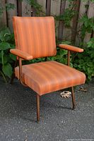 Front angled view of the vintage armchair showing orange striped vinyl upholstery, padded seat and back, and metal frame with armrests.