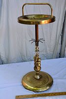 Full view of the vintage metal ashtray stand showing the handle, tray, column with cherub figures, and round base.